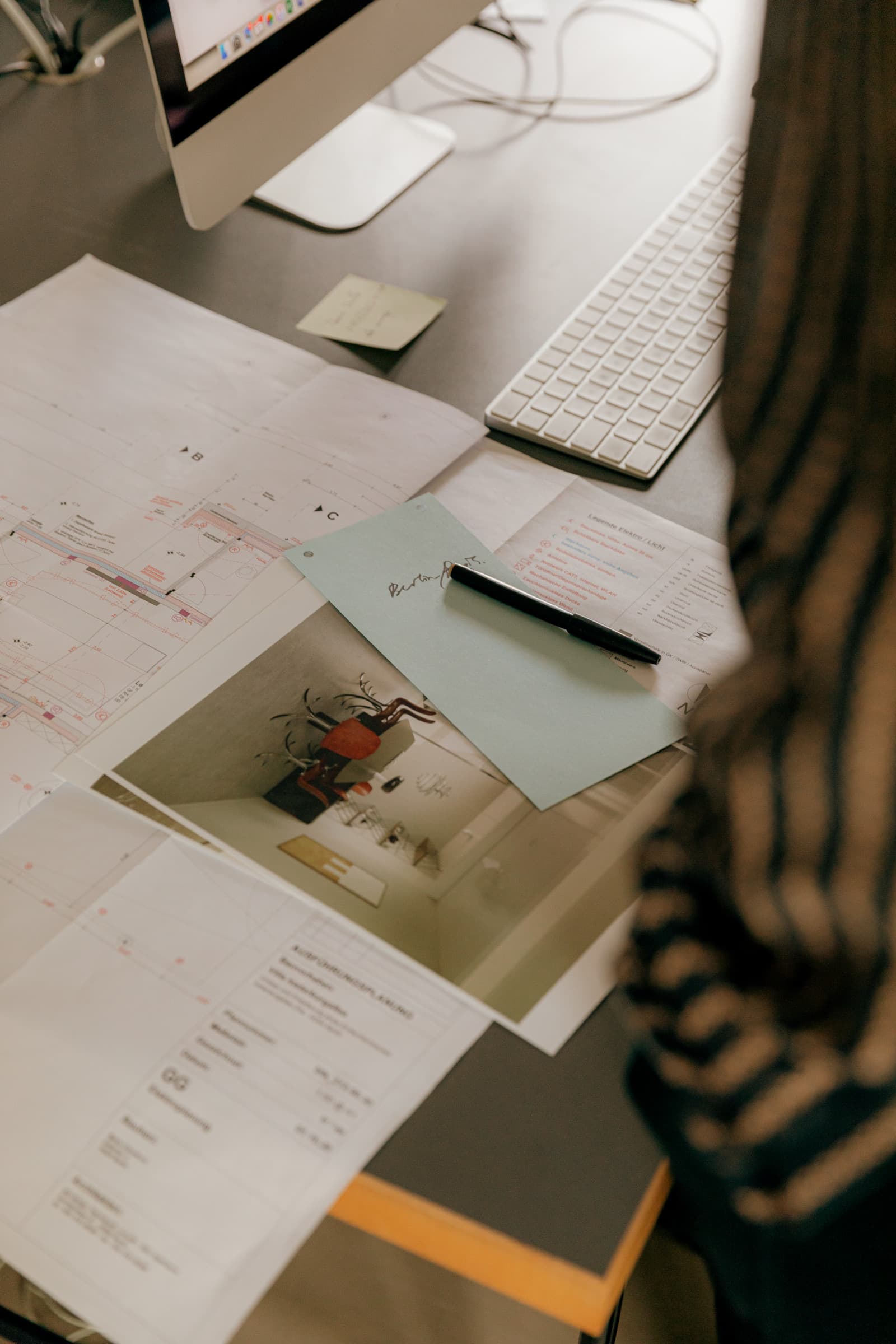 Grundrisse, gedrucktes Innenraumfoto und Stift verteilt auf dem Schreibtisch neben der Tastatur, über die Schulter einer Person gesehen