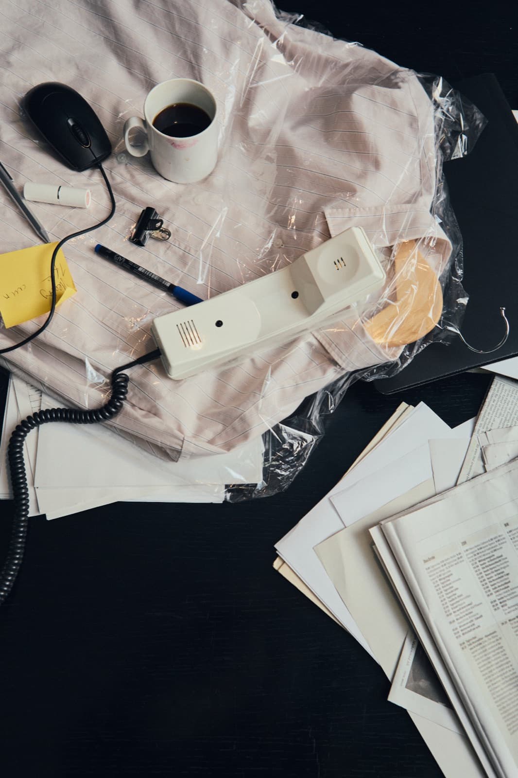 Vollgestellter Schreibtisch von oben: Telefonhörer mit Kabel, Kaffeebecher und Maus auf folienverpacktem Hemd neben losen Blättern