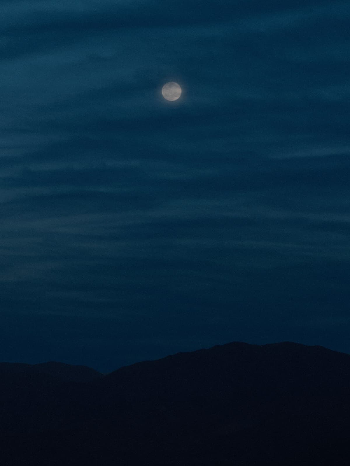 Vollmond leuchtet durch dünne Wolkenstreifen, hoch über der Silhouette eines Bergkamms