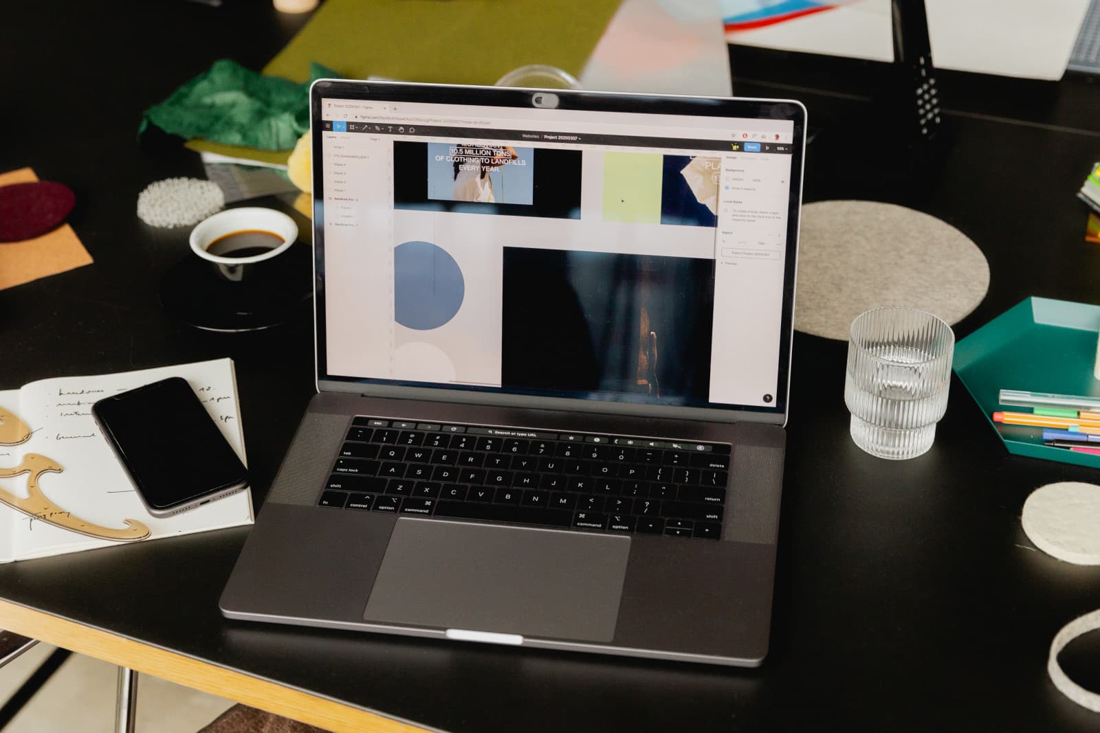 Offener Laptop zeigt ein Designprogramm mit Bildkacheln, daneben handbeschriebenes Notizbuch, Handy, Kaffeetasse und Wasserglas